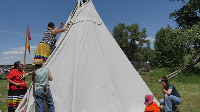 Indigenous peoples in ribbon skirts, a Parks Canada staff member, and some visitors help set up a tipi. 