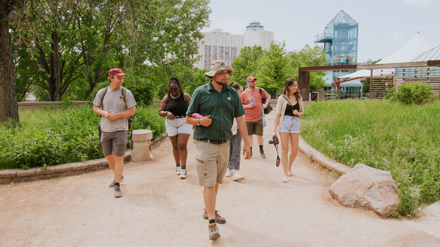 A guide in a Parks Canada uniform gives a tour to 5 visitors at The Forks on a summer day. 