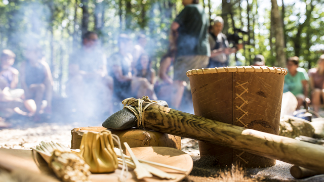 A closeup shot of indigenous instruments and tools. Behind them and out of focus, an elder wearing a Parks Canada uniform speaks to a crowd of people near a bonfire.