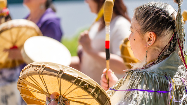 An Indigenous woman in cultural regalia leads a drumming circle.