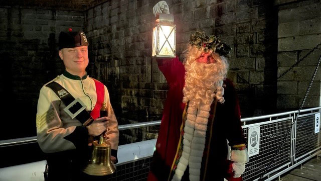 A man in uniform next to a man in a Santa Claus costume holding a lantern with the Halifax Citadel in the background. 