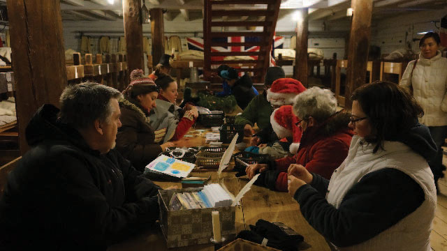 A group of visitors sit around a table in winter coats and santa costumes as they  partake in christmas crafts.