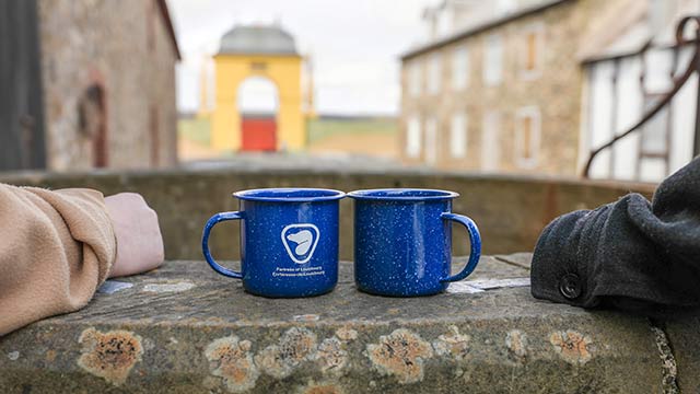 Two Parks Canada camping mugs sitting on a rusted ledge with historic buildings in the background. 