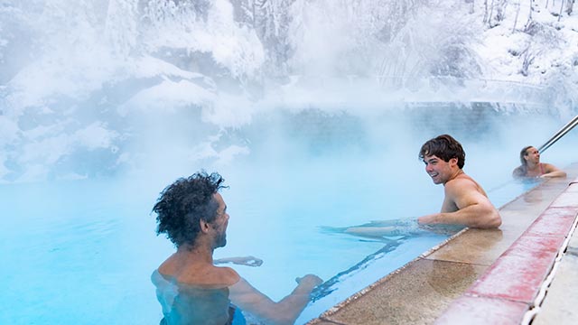 Three people in a pool at Radium Hot Springs, with steam rising and snow-covered trees in the background.