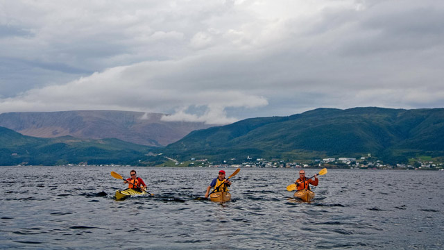 Three visitors paddle on sea kayaks on Bonne Bay at Norris Point on a cloudy day in Gros Morne National Park.