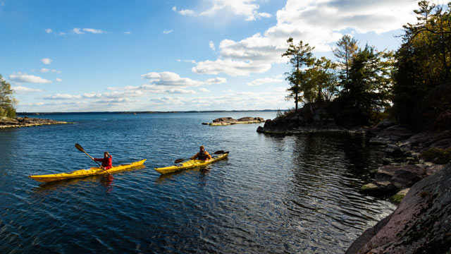 Two visitors kayak on the St. Lawrence River near a rocky shore in Thousand Islands.