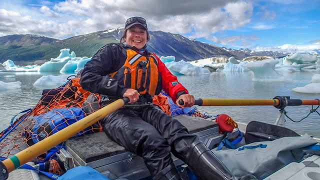 A woman smiles while rafting on the Alsek River with mountains and small floating ice blocks in the background in Kluane.