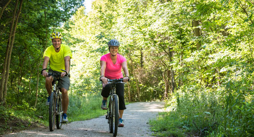 Deux cyclistes profitent de la piste cyclable du Centenaire bordée d'arbres au feuillage vert.