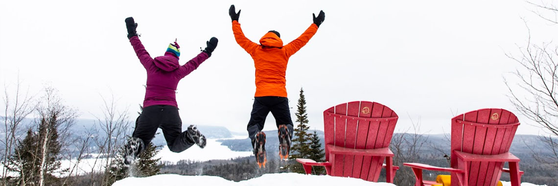 Two hikers wearing crampons jump near the red chairs at the Lac-Rosoy lookout in La Mauricie National Park.