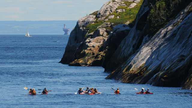 Kayakers in the Saguenay–St. Lawrence Marine Park with cliffs, a boat and a lighthouse in the background.