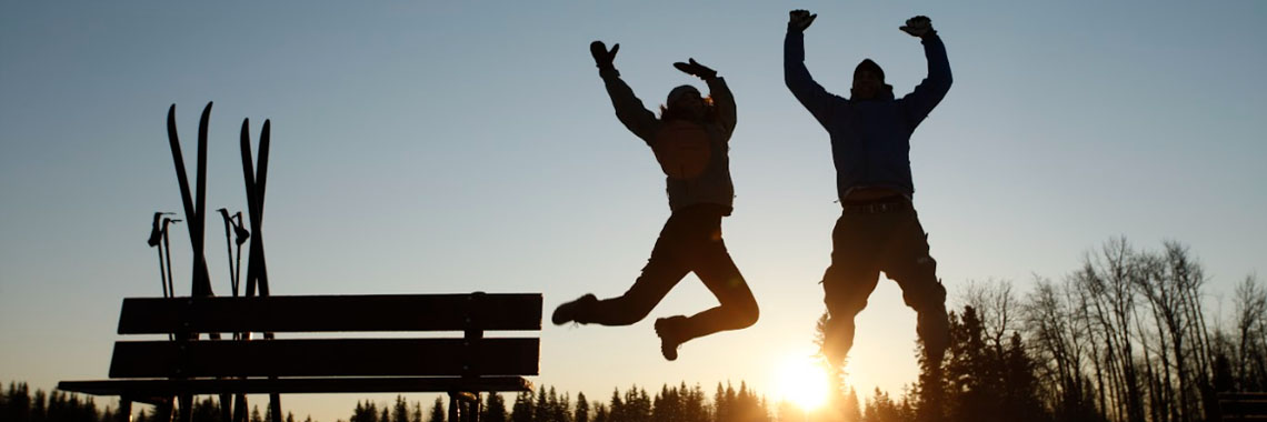 Two people jump for joy near a bench where their skis are leaning during sunset at Elk Island National Park.
