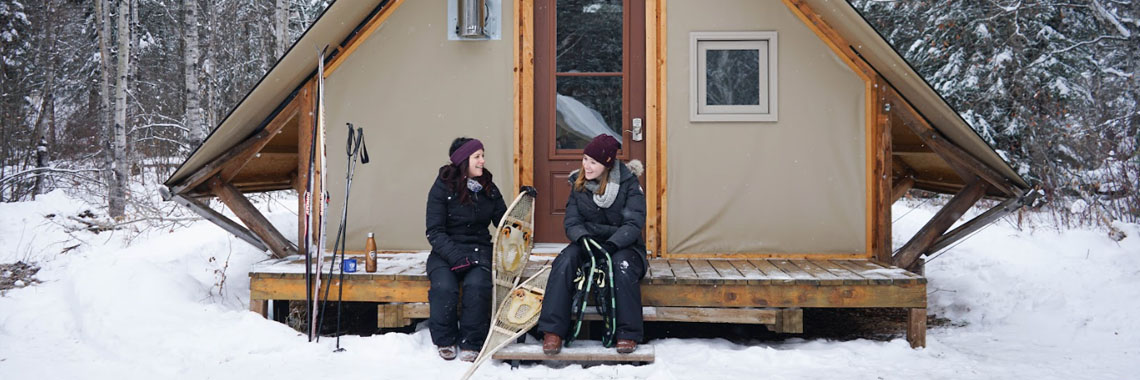 Two young women sitting on the deck of their oTENTik tent in winter at Riding Mountain National Park.