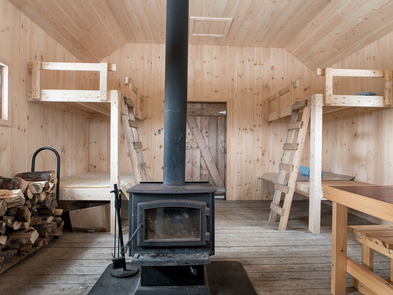 The interior of a cabin: a wooden table, benches, two bunk beds, a counter, a wood stove and a pile of firewood. Fundy National Park's Hastings rustic cabin