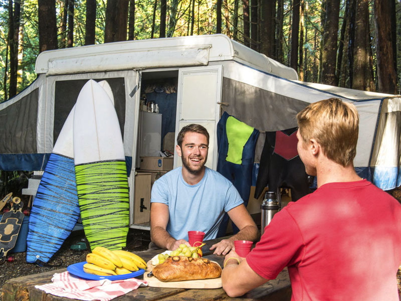 Two young adults share a meal near their tent-trailer on which two surfboards are leaning.