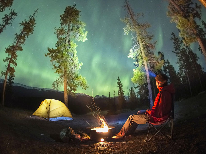 A man sits near a campfire beside his tent and looks at the northern lights.