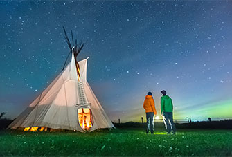 Two visitors gaze at the Big Dipper outside a tipi on a starry night.