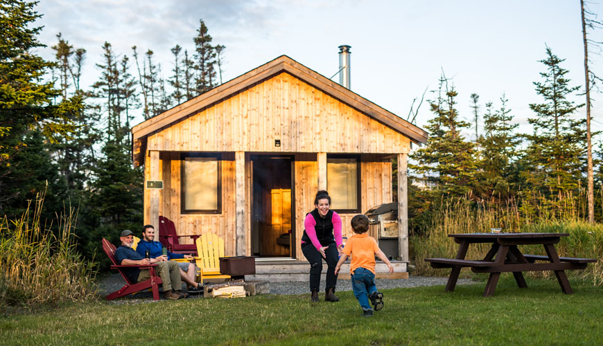 A child runs to his mother as two men sit in front of a rustic wooden cabin.