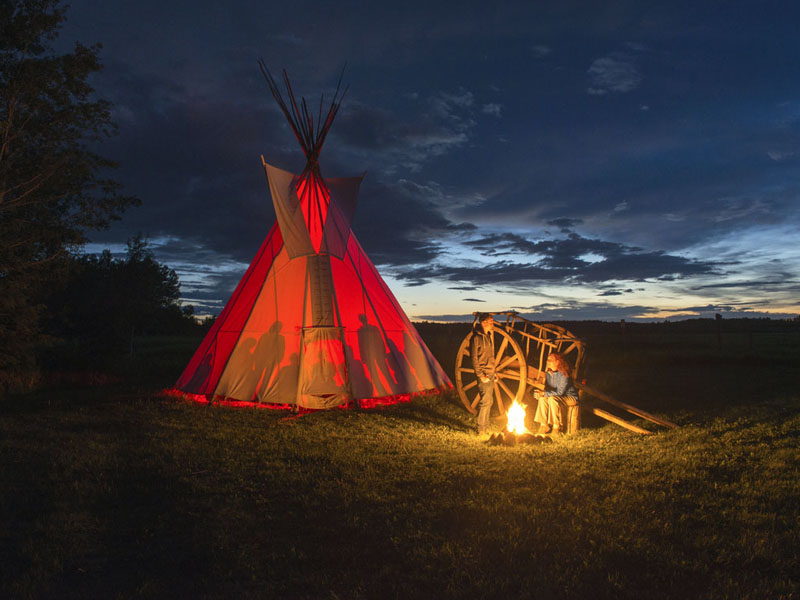 Two people at a campfire beside a Red River cart and a glowing red tipi at dusk.