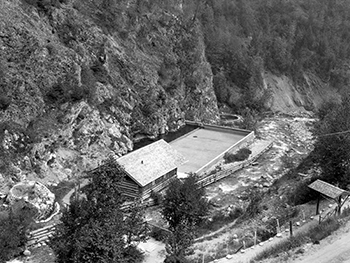 Vue plongeante de la piscine en béton, du vestiaire en rondins et du sentier menant aux sources thermales Radium