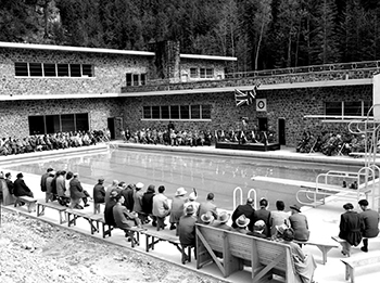 Une foule assise sur des bancs autour de la piscine d’eau froide des sources thermales Radium. Un orateur est sur scène