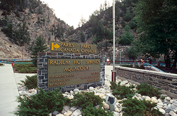 Stone base and brown wooden sign with yellow lettering. Sign says Parks Canada, Radium Hot Springs Aquacourt, Swimming pools, information, and restaurant in French and English