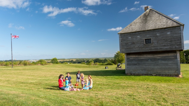 At a distance, a family sit on a blanket picnicking on the lawn near the wooden blockhouse.