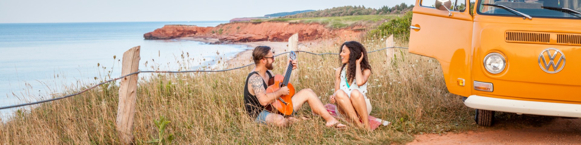 Two adults sitting in the grass by the ocean next to a Westfalia.