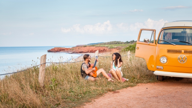 Two adults sitting in the grass by the ocean next to a Westfalia.