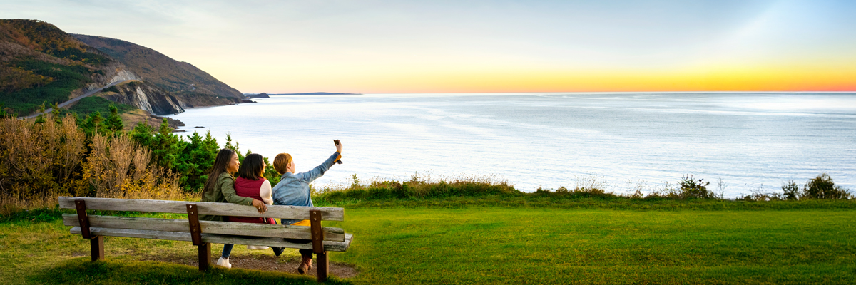 Three visitors take a selfie on a bench along the Cabot Trail with the ocean in the background.