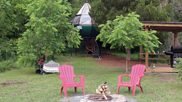 A teardrop-shaped Ôasis accommodation on a campground with a barbecue, two red chairs and a fire pit.