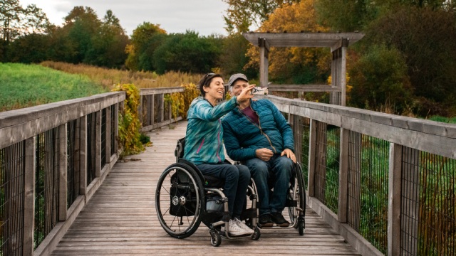 Two adults in wheelchairs take a selfie on a boardwalk.