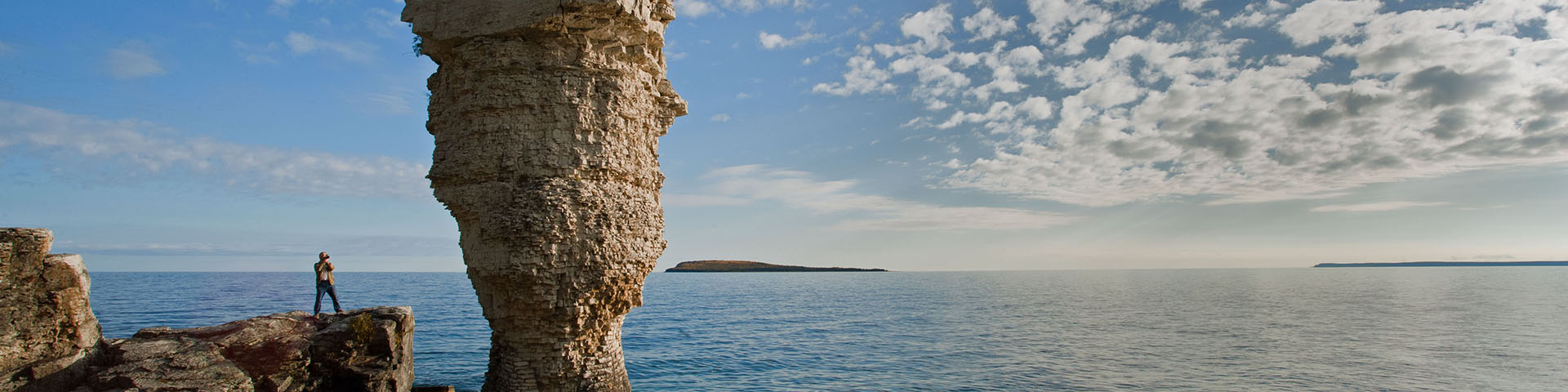 An adult photographs a flowerpot-shaped rock on Flowerpot Island with Lake Huron in the background at Fathom Five National Marine Park.