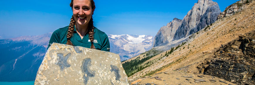 Une guide de Parcs Canada tient un fossile à la carrière Walcott, avec une vue du lac Emerald, du glacier Emerald et du sommet du mont Wapta.