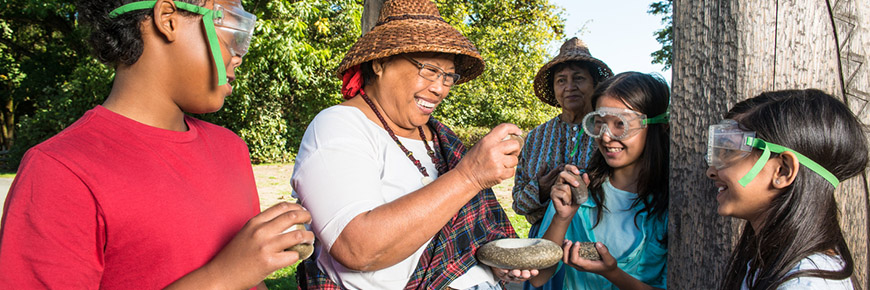 A First Nation interpreter encourages a group of children to try “Pecking Stones” to create stone weights for fishing nets