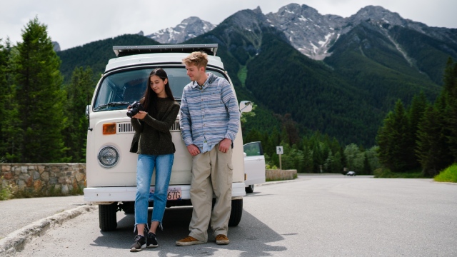 Two visitors with a camera in front of their Westfalia, mountains in the background.