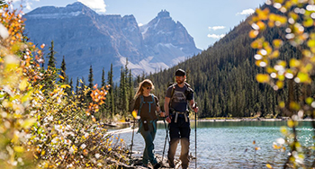 Two visitors hike along the edge of a lake with mountains in the background in Yoho.