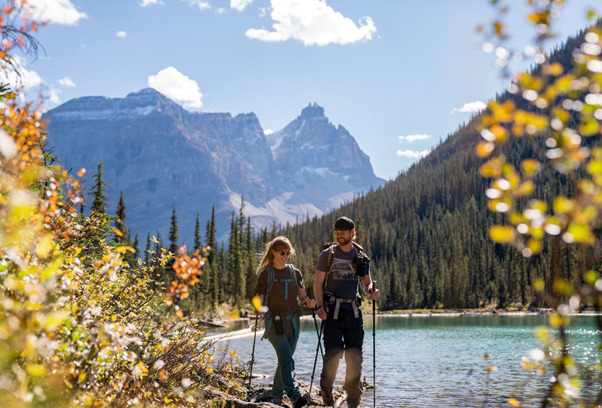 Two visitors hike along the edge of a lake with mountains in the background in Yoho.