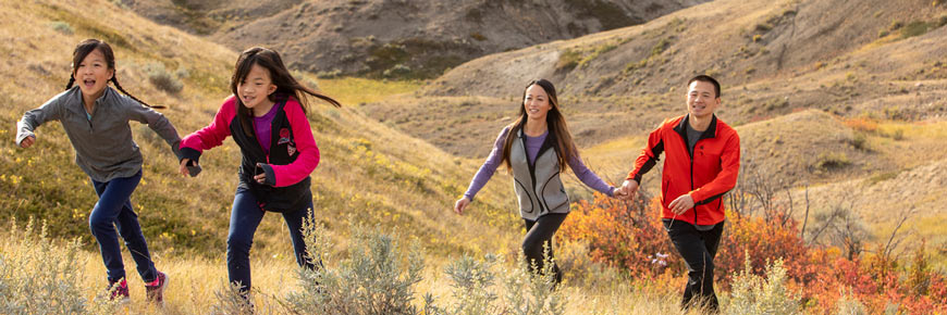 Two children running up a hill, with their parents following, with the landscape of the West Block of Grasslands National Park in the background.