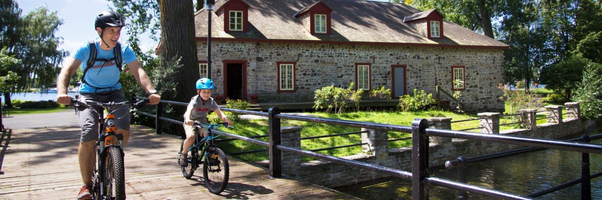 An adult and a child biking on the canal’s path with a stone building in the background.