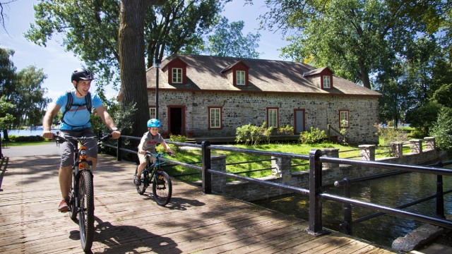 Un adulte et un enfant en vélo sur la piste du canal avec un bâtiment en pierre en arrière-plan.