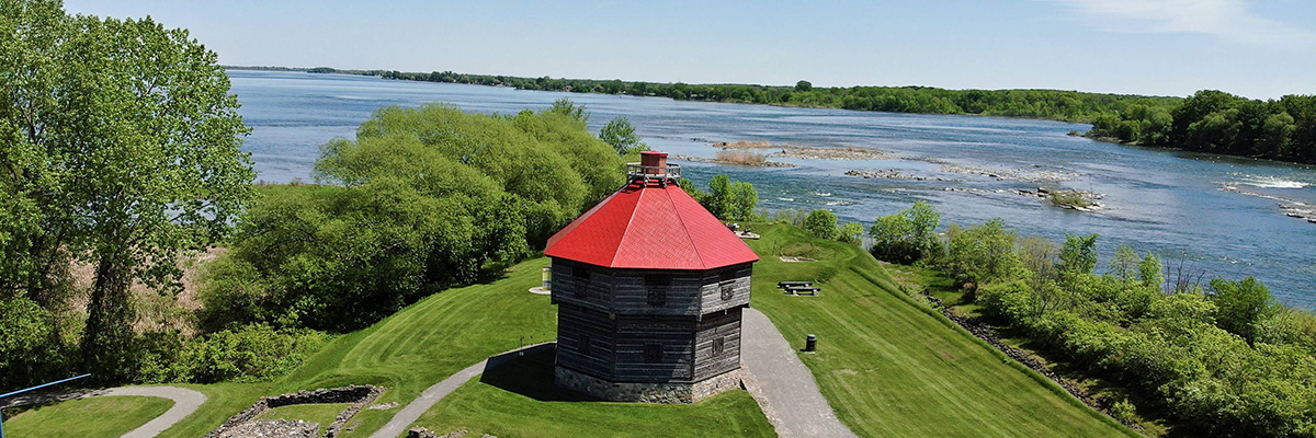 Un blockhaus avec un toit rouge et des vestiges en bordure du fleuve à Coteau-du-Lac.