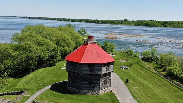 Un blockhaus avec un toit rouge et des vestiges en bordure du fleuve à Coteau-du-Lac.