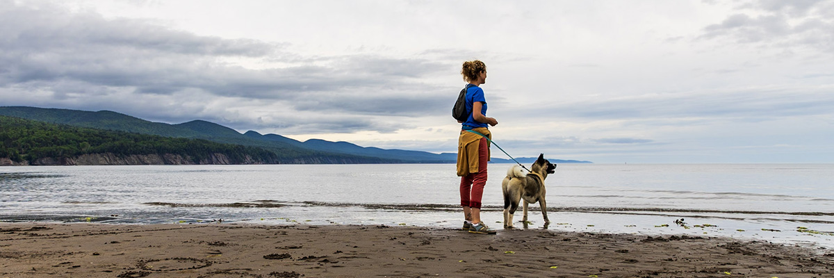A woman and her leashed dog on Penouille Beach with Gaspé Bay in the background in Forillon.