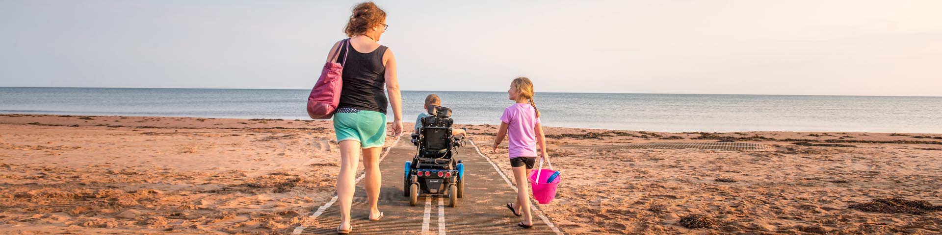 Une famille présentant des besoins en matière d’accessibilité marche en direction de l’eau sur la plage Stanhope en utilisant des tapis de mobilité.