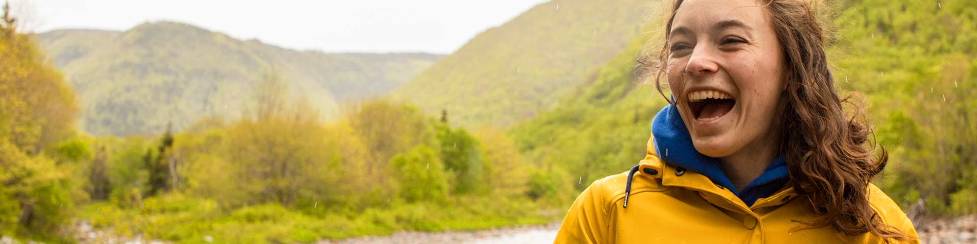 Une femme souriante portant un manteau de pluie jaune à l’extérieur, avec des montagnes verdoyantes et une légère pluie en arrière-plan.