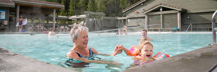 A grandmother and her grandchild playing in Miette Hot Springs on a sunny day, surrounded by large green pine trees in the back drop.  