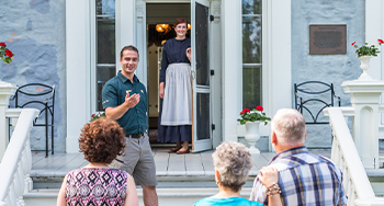 Un jeune guide de Parcs Canada ainsi qu’une guide en costume d’époque accueillent un groupe de visiteurs sur le parvis du chic manoir au lieu historique national du Manoir-Papineau.
