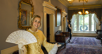 Une jeune guide de Parcs Canada vêtue d’une chic robe d’époque et munie d’un éventail sourit et accueille des visiteurs dans le vestibule du lieu historique national du Manoir-Papineau.