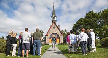 Un interprète de Parcs Canada guide des visiteurs lors d'une visite au lieu historique national de Grand-Pré, avec l'église-souvenir en arrière-plan.