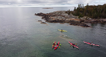 Un groupe de kayakistes pagayant près d'un rivage rocheux avec des arbres, sous un ciel nuageux sur des eaux calmes.